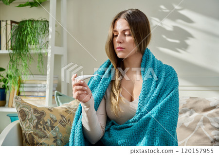Young woman using thermometer while sitting with blanket on bed at home Young woman using thermometer while sitting with blanket on bed at home 120157955
