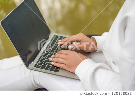 A woman is typing on a laptop while sitting on a chair. She is wearing a white shirt and white pants. The laptop is open and the screen is black. The woman is focused on her work. 120158021