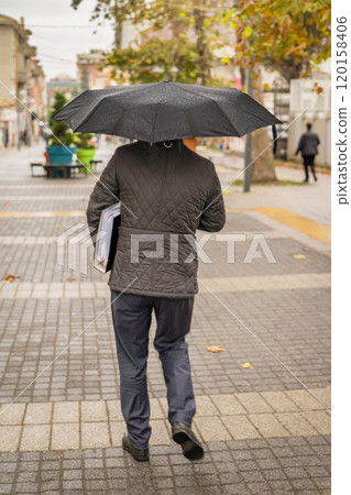 Rear view of man with a black umbrella walking down wet city street holding documents underarm 120158406
