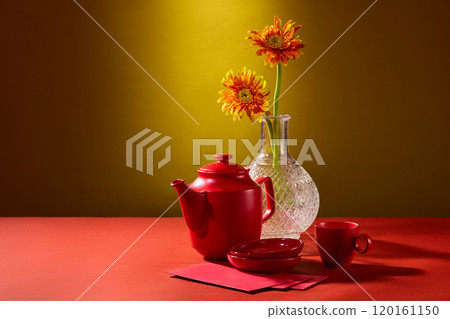 A modern Tet-themed shows a table with glass vases containing gerbera flowers, a tea set, and lucky envelopes resting on the red table is seen in close-up against a yellow backdrop. 120161150