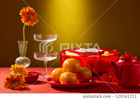 A close-up of a contemporary Tet banquet table with gerbera flowers, glasses, teapot, jam tray, and tangerines dish surrounded by a red tabletop, depicted in a straw-colored backdrop, 120161151