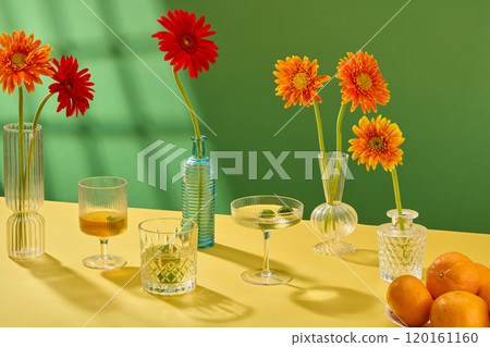 A modern Tet concept in the still life photo template, front view of water glasses placed next to flower vases arranged with gerbera, and tangerines dish on a yellow table, in the green background. 120161160