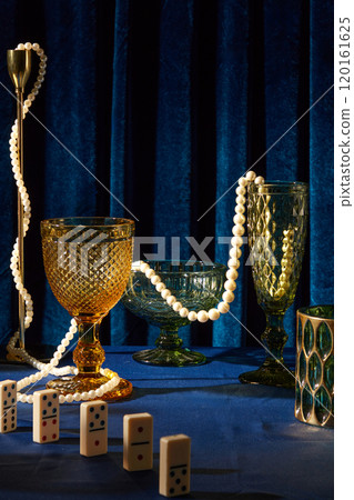 Glasses with different shapes and colors are placed on blue tabletop, surrounded by brass candle holders, a pearl necklace, and dominoes. New Year Eve theme close up image on a ultramarine backdrop. 120161625
