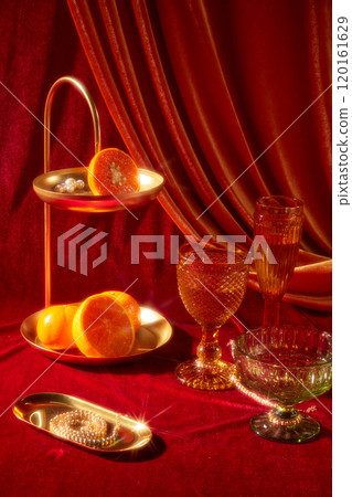 A close-up New Years Eve party idea image has a variety of glasses next to a brass tray, oranges, pearls, and a long copper tray with a pearl necklace on a red surface against a crimson backdrop. 120161629