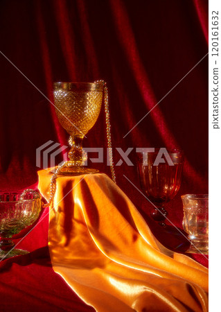 A glass cup wrapped in gold jewelry sits on a platform covered with a gold and red fabric. On the red surface are glasses of different shapes and colors. Close-up image of the New Years Eve content. 120161632