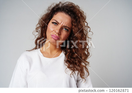 Woman with curly hair expresses confusion in a studio setting against a neutral background 120162871