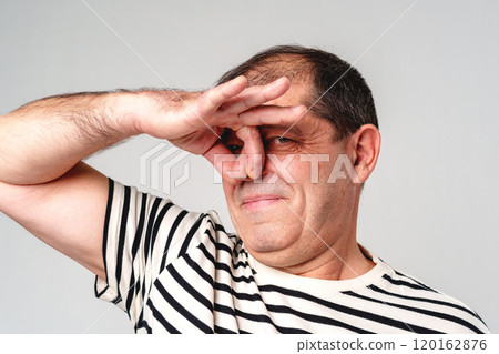 Middle-aged man playfully pinching his nose with a smile in a striped shirt against a plain background Middle-aged man playfully pinching his nose with a smile in a striped shirt against a plain background 120162876
