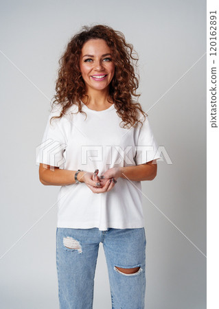 A young woman with curly hair wearing a white t-shirt and ripped jeans stands confidently against a gray backdrop 120162891