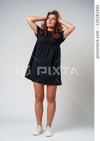 Smiling woman with curly hair poses confidently against a plain background while holding her hands behind her head 120162892