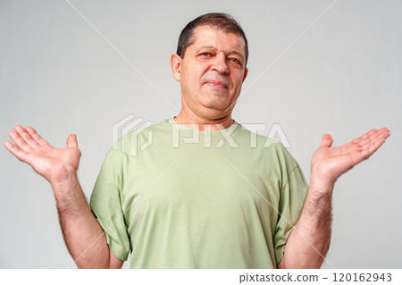 A middle-aged man with a neutral expression raises his hands in a questioning gesture against a plain backdrop 120162943