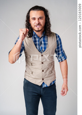 A young man with curly hair dressed in a vest and plaid shirt shows determination while making a fist indoors 120163009