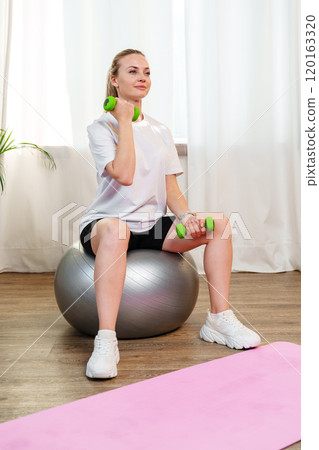 Woman exercising with dumbbells on a stability ball in a indoor space Woman exercising with dumbbells on a stability ball in a indoor space 120163320