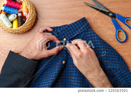 A person sewing a button on a blue plaid shirt with sewing tools and threads on a wooden table 120163501
