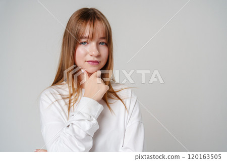 Young woman in a white long-sleeve shirt thoughtfully poses with a hand on her chin in a minimalistic setting 120163505
