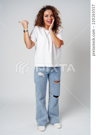 A young woman with curly hair playfully pointing finger while standing against a gray background 120163557
