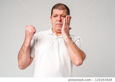 An older man holds his cheek, showing concern, against a plain background An older man holds his cheek, showing concern, against a plain background 120163684