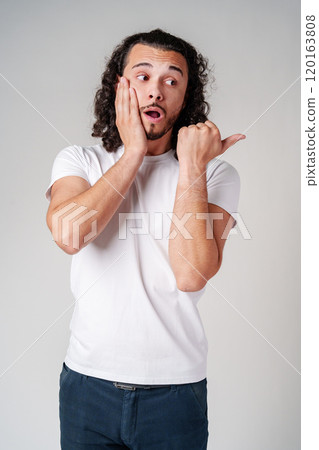 Young man in a white t-shirt and jeans pointing confidently while smiling in a studio setting 120163808