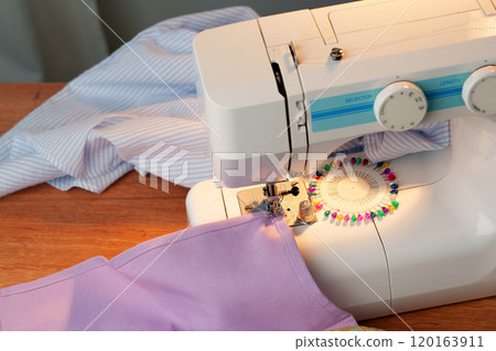 Sewing machine in use on a wooden table, stitching a light fabric in a well-lit indoor workspace Sewing machine in use on a wooden table, stitching a light fabric in a well-lit indoor workspace 120163911