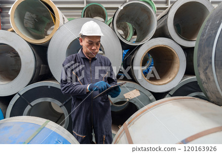 A worker checks a tablet in front of large rolls of metal. Safety gear is worn for protection in the workplace. 120163926