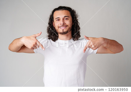 A young man with curly hair joyfully pointing down while wearing a plain white t-shirt against a neutral background 120163950