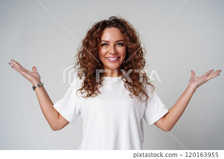 Cheerful woman with curly hair poses with open arms in a white shirt against a plain background Cheerful woman with curly hair poses with open arms in a white shirt against a plain background 120163955