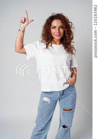 A young woman with curly hair playfully pointing finger while standing against a gray background 120163962