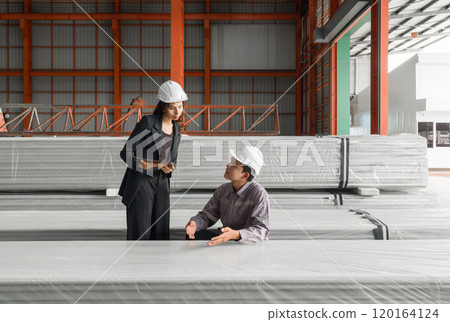 Two worker in hardhat talk in a warehouse. They discuss plans while standing near large, wrapped material. 120164124