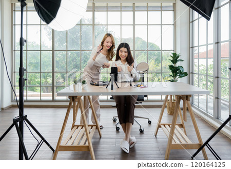 Two asian women creating content for social media. They are using a studio setup that include a smartphone mounted on a tripod, lighting equipment and various makeup products arrayed on the table. Two asian women creating content for social media. They are using a studio setup that include a smartphone mounted on a tripod, lighting equipment and various makeup products arrayed on the table. 120164125