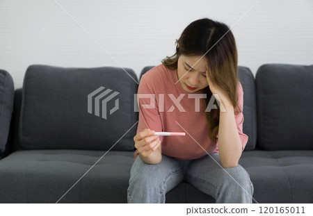 Young asian teen stressed depressed sitting on sofa in living room holding positive result fertilization pregnancy test kit. 120165011