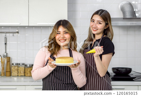Two young women in apron make dessert in the kitchen. One people holding a piping bag. The other holding cake on white dish. Two young women in apron make dessert in the kitchen. One people holding a piping bag. The other holding cake on white dish. 120165015