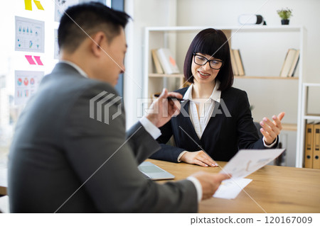 Asian businessman presents report to Caucasian businesswoman at office 120167009