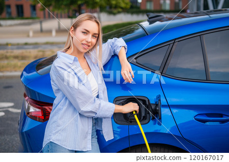 Portrait of blonde woman in casual clothes charging her electric car at a public parking station 120167017