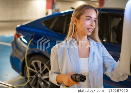 Blonde woman selects charging mode on the charging station on the parking. Blonde woman selects charging mode on the charging station on the parking. 120167020