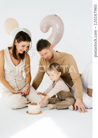 Young family with a child is sitting on the floor in front of a birthday cake in honor of the second birthday with balloons and the number two Young family with a child is sitting on the floor in front of a birthday cake in honor of the second birthday with balloons and the number two 120167960