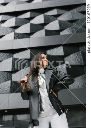 Young woman wearing a black jacket and white pants stands in front of a building. She is smiling and she is enjoying herself 120167969