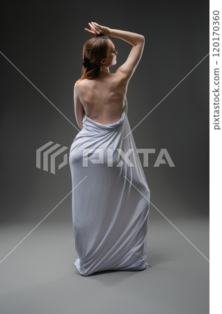 Elegant woman draped in light fabric poses gracefully against a dark backdrop 120170360