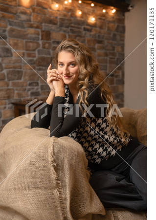 Young woman posing with a smile on a cozy couch in a warm, rustic living room setting Young woman posing with a smile on a cozy couch in a warm, rustic living room setting 120171135