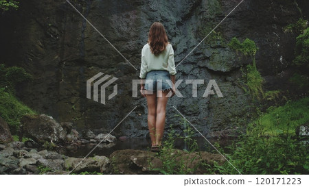 A young woman in a white shirt and denim shorts stands on a rocky outcrop near a waterfall in a lush, tropical forest in French Polynesia. The waterfall cascades down a dark, mossy rock face. A young woman in a white shirt and denim shorts stands on a rocky outcrop near a waterfall in a lush, tropical forest in French Polynesia. The waterfall cascades down a dark, mossy rock face. 120171223