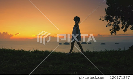 Woman gracefully walks grass hill, sunset sky in background. Female enjoy aerial view on tropical island. Girl silhouette against colorful orange sky. Outdoor lifestyle travel, summer holiday vacation Woman gracefully walks grass hill, sunset sky in background. Female enjoy aerial view on tropical island. Girl silhouette against colorful orange sky. Outdoor lifestyle travel, summer holiday vacation 120171277