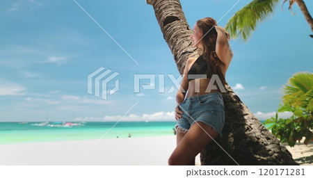 Young woman is enjoying her tropical vacation, relaxing on a white sand beach, leaning against a palm tree, with turquoise water and blue sky in the background Young woman is enjoying her tropical vacation, relaxing on a white sand beach, leaning against a palm tree, with turquoise water and blue sky in the background 120171281