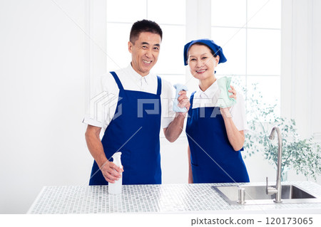 Senior housekeepers (male and female, couple) wearing blue aprons and white polo shirts standing side by side in the kitchen 120173065