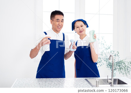 Senior housekeepers (male and female, couple) wearing blue aprons and white polo shirts standing side by side in the kitchen Senior housekeepers (male and female, couple) wearing blue aprons and white polo shirts standing side by side in the kitchen 120173066