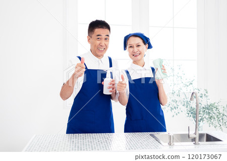 Senior housekeepers (male and female, couple) wearing blue aprons and white polo shirts standing side by side in the kitchen Senior housekeepers (male and female, couple) wearing blue aprons and white polo shirts standing side by side in the kitchen 120173067