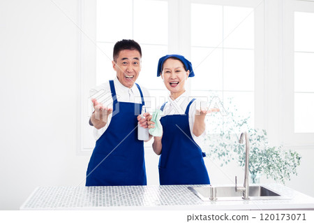Senior housekeepers (male and female, couple) wearing blue aprons and white polo shirts standing side by side in the kitchen 120173071
