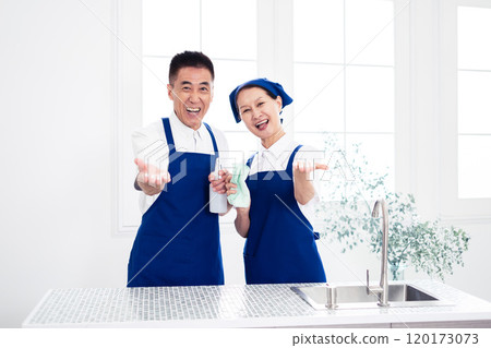 Senior housekeepers (male and female, couple) wearing blue aprons and white polo shirts standing side by side in the kitchen 120173073
