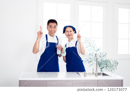 Senior housekeepers (male and female, couple) wearing blue aprons and white polo shirts standing side by side in the kitchen 120173077