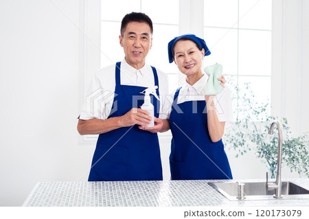 Senior housekeepers (male and female, couple) wearing blue aprons and white polo shirts standing side by side in the kitchen 120173079