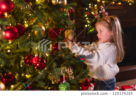 Merry Christmas Xmas holiday. Little girl decorating Christmas tree on Christmas eve at home. Young kid near classical Christmas tree with winter decorations ornaments. Time for celebration Christmas 120174285