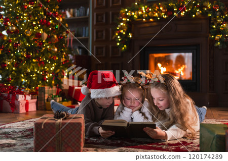 Merry Christmas Xmas holiday. Brother sisters children kids reading magic Christmas story book near Christmas tree in living room with fireplace in winter evening. Christmas eve at home Merry Christmas Xmas holiday. Brother sisters children kids reading magic Christmas story book near Christmas tree in living room with fireplace in winter evening. Christmas eve at home 120174289