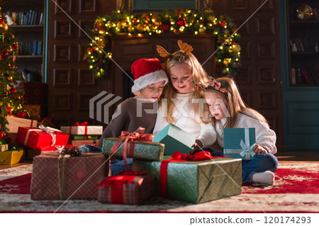 Merry Christmas Xmas holiday. Children unpacking open up gift box near Christmas tree. Kids in living room with Christmas tree fireplace opening gift box with surprise faces. Christmas eve at home Merry Christmas Xmas holiday. Children unpacking open up gift box near Christmas tree. Kids in living room with Christmas tree fireplace opening gift box with surprise faces. Christmas eve at home 120174293